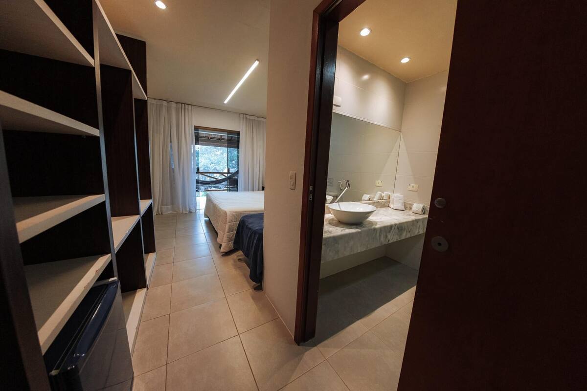 Habitación de hotel moderna con cama, baño con espejo grande y lavamanos, y vista al exterior por una ventana.