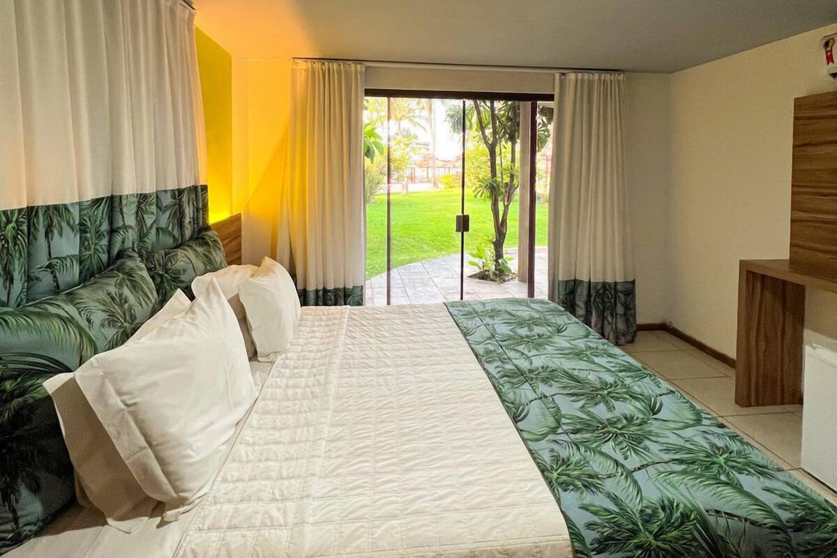 Un dormitorio con una cama grande, cortinas, vistas a un jardín y una mesita de noche de madera.