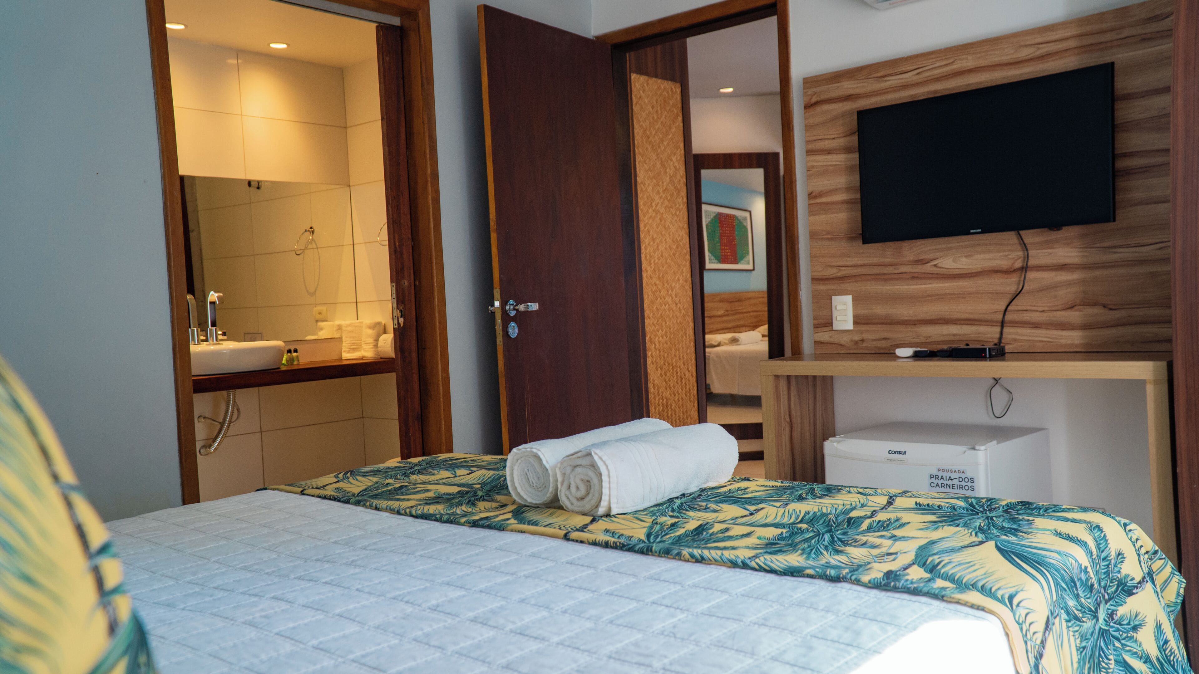 A hotel room with a bed, a television mounted on a wooden panel, a bathroom with a sink and mirror, and a door leading to another room.