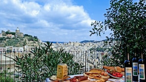 Una mesa con varios quesos, embutidos y pan, con un paisaje urbano y una colina con una estructura en forma de cúpula de fondo.