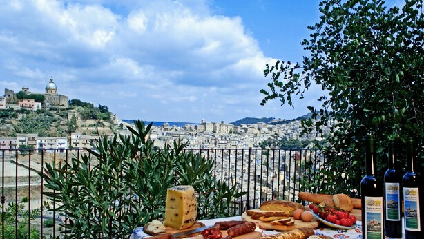 Una mesa con varios quesos, embutidos y pan, con un paisaje urbano y una colina con una estructura en forma de cúpula de fondo.