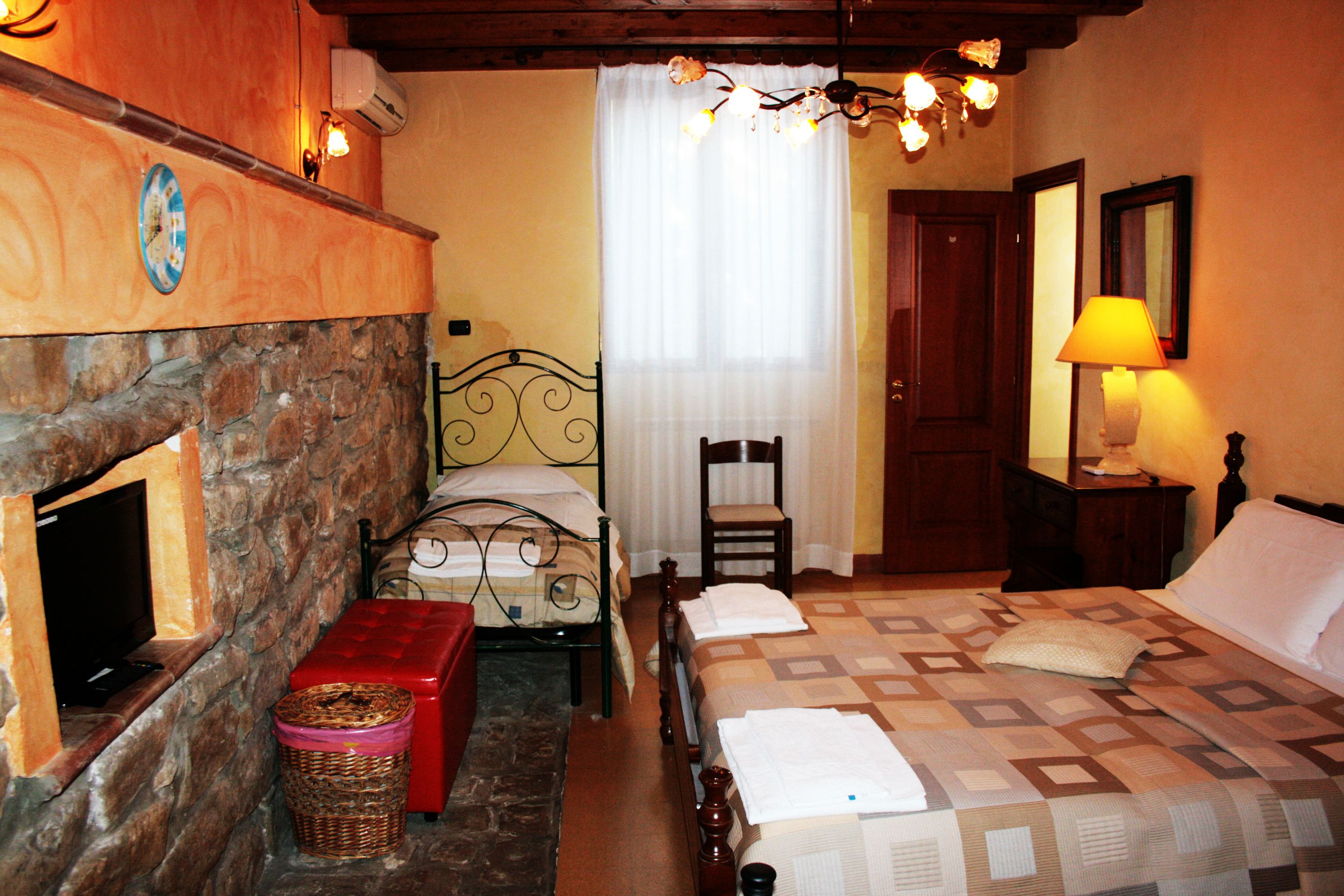 Un dormitorio con chimenea de piedra, una cama con colcha estampada, una silla, una mesita de noche de madera con lámpara y un espejo en la pared.