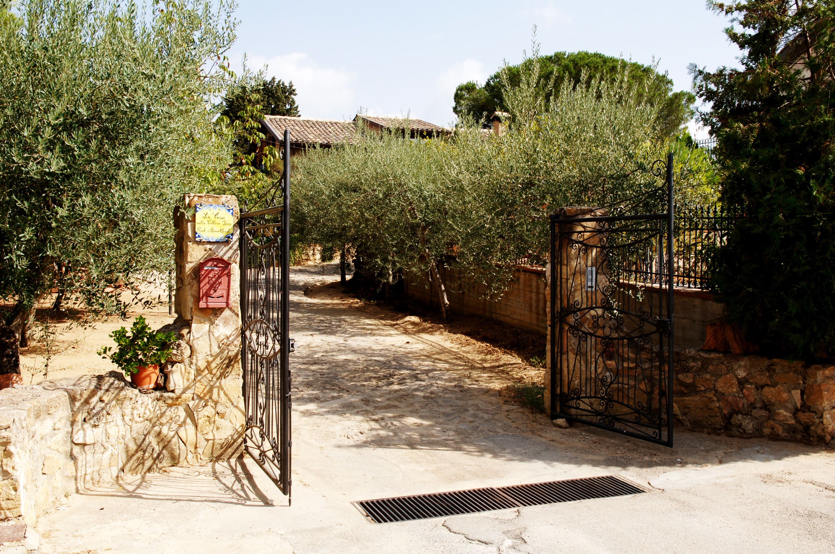Una entrada protegida por un muro de piedra y una reja metálica, que conduce a un sendero bordeado de olivos y un edificio con tejado de tejas al fondo.