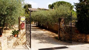 Una entrada protegida por un muro de piedra y una reja metálica, que conduce a un sendero bordeado de olivos y un edificio con tejado de tejas al fondo.