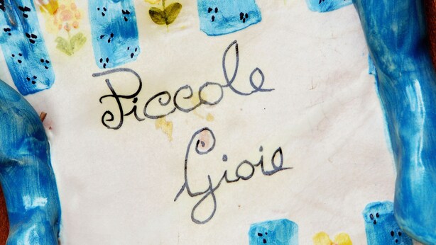 Un trozo de papel con el nombre "Piccole Gioie" escrito, rodeado de acuarelas en azul y amarillo de flores y hojas.