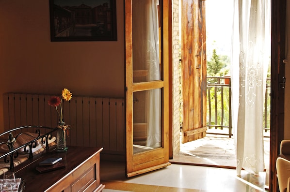 A room with a wooden door leading to a balcony, a bed with a headboard, a bedside table with a vase of flowers, and a framed picture on the wall.