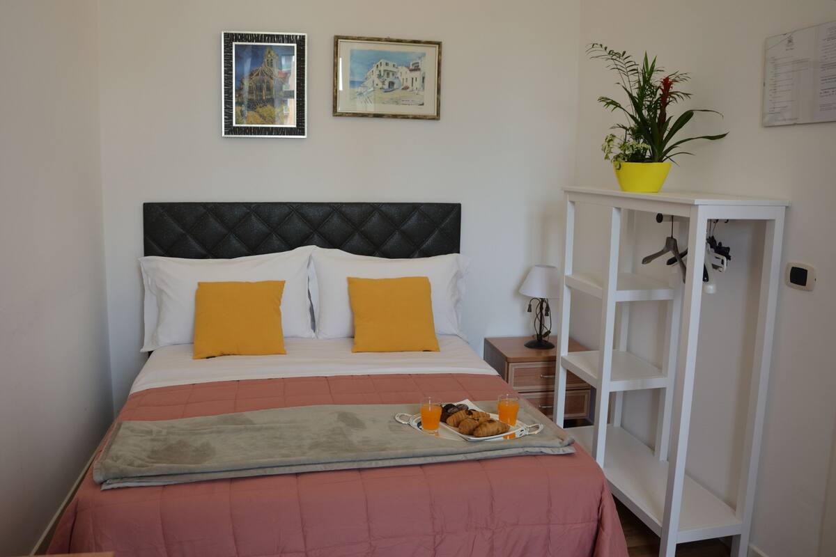 Un lit bien fait, avec une tête de lit matelassée, deux oreillers, un plateau avec des croissants et du jus d’orange, et une plante en pot posée sur une commode blanche.