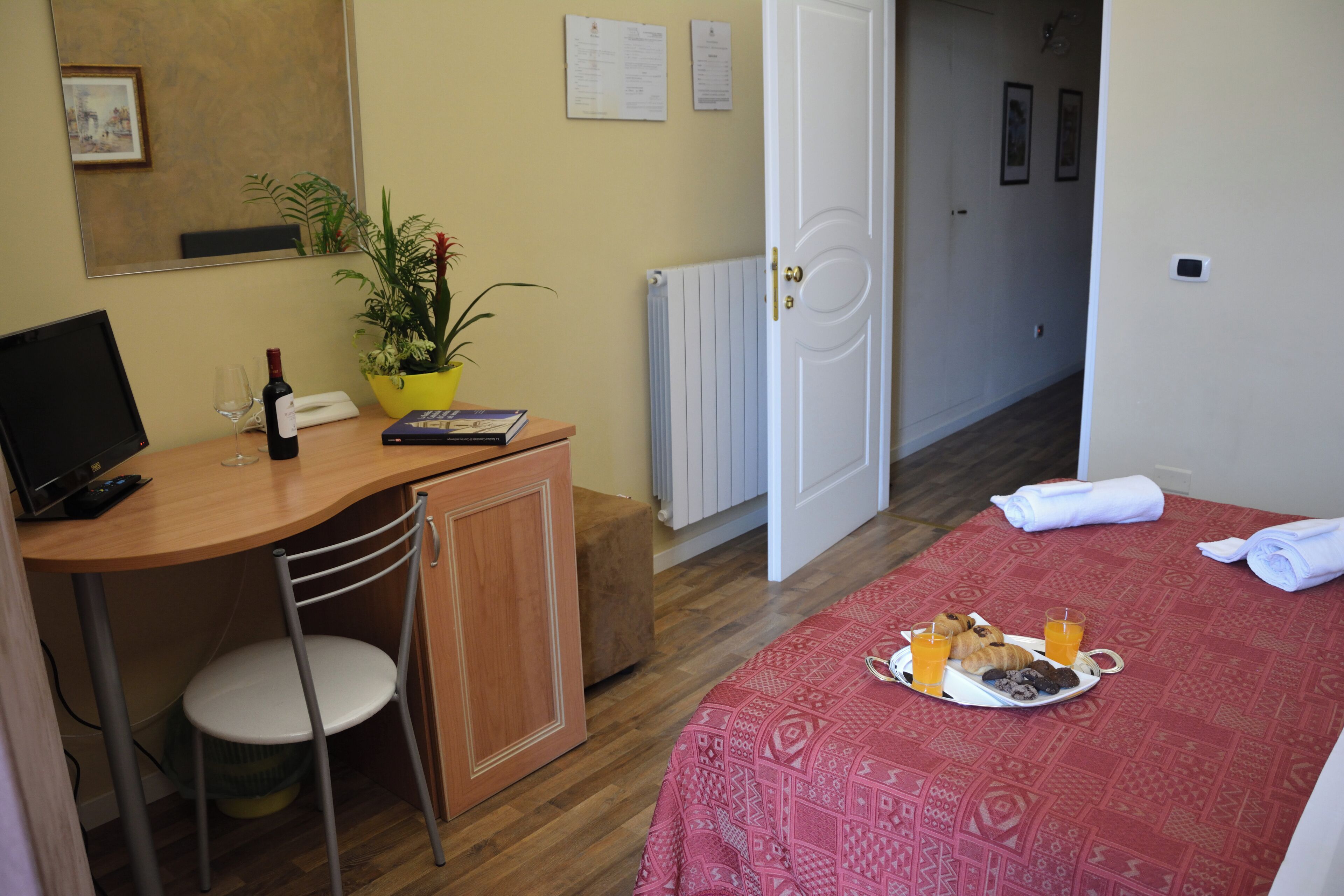 A hotel room with a wooden desk, a chair, a bed with a red patterned bedspread, a tray with breakfast items, a potted plant, and a radiator.