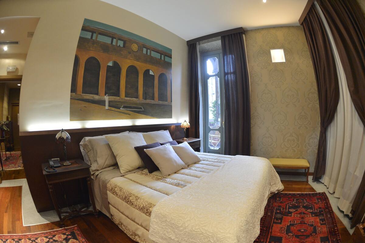 Un dormitorio ordenado con una cama grande, una mesita de noche, un banco y un cuadro de un puente en la pared.