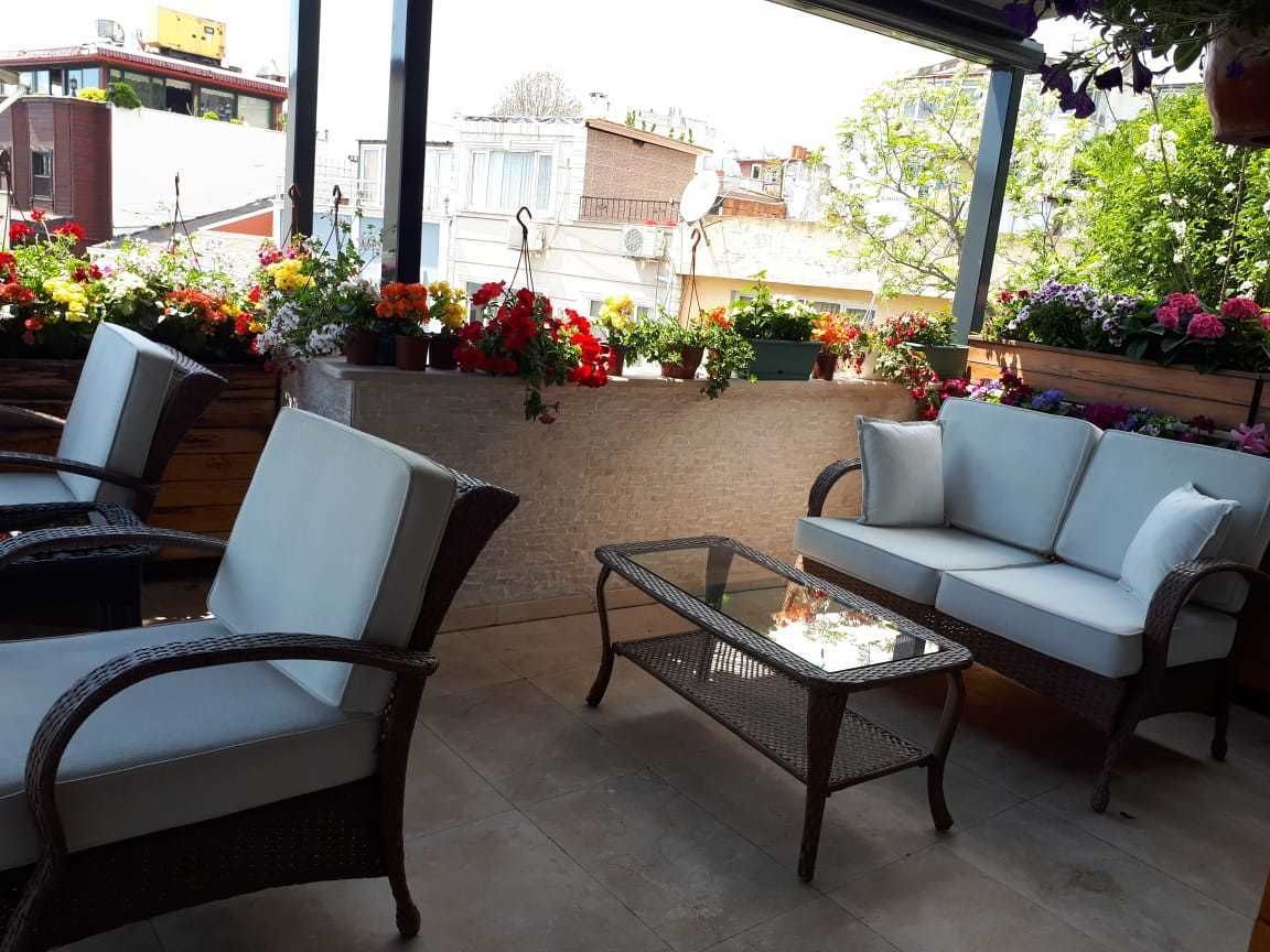 Un área de estar al aire libre con sillas de mimbre y una mesa de centro de vidrio, rodeada de plantas y flores en macetas.