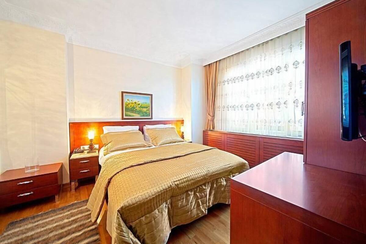A hotel room with a large bed, a wooden nightstand, a desk, and a window with curtains.