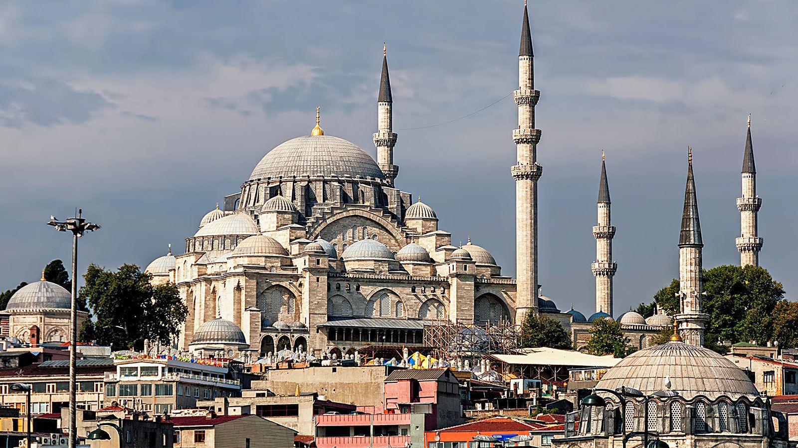 A historic mosque with multiple domes and minarets, surrounded by urban buildings and a busy street.