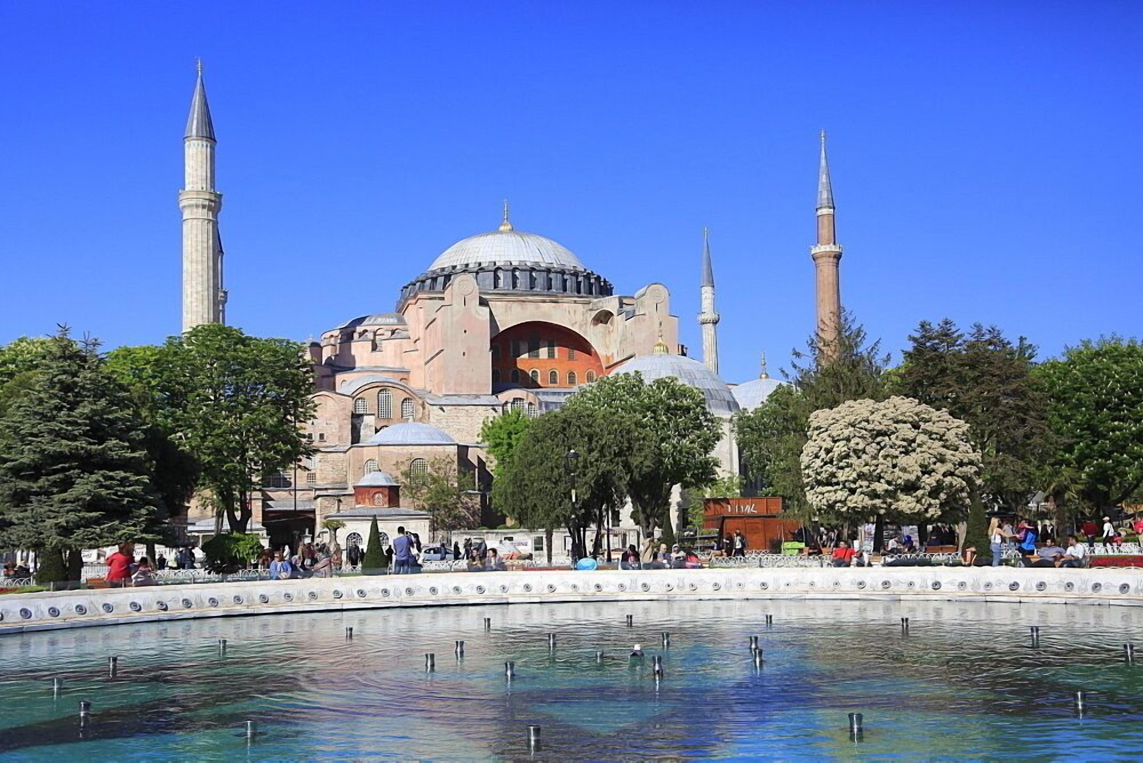 A historic building with multiple minarets and a large central dome, surrounded by a water feature and trees.