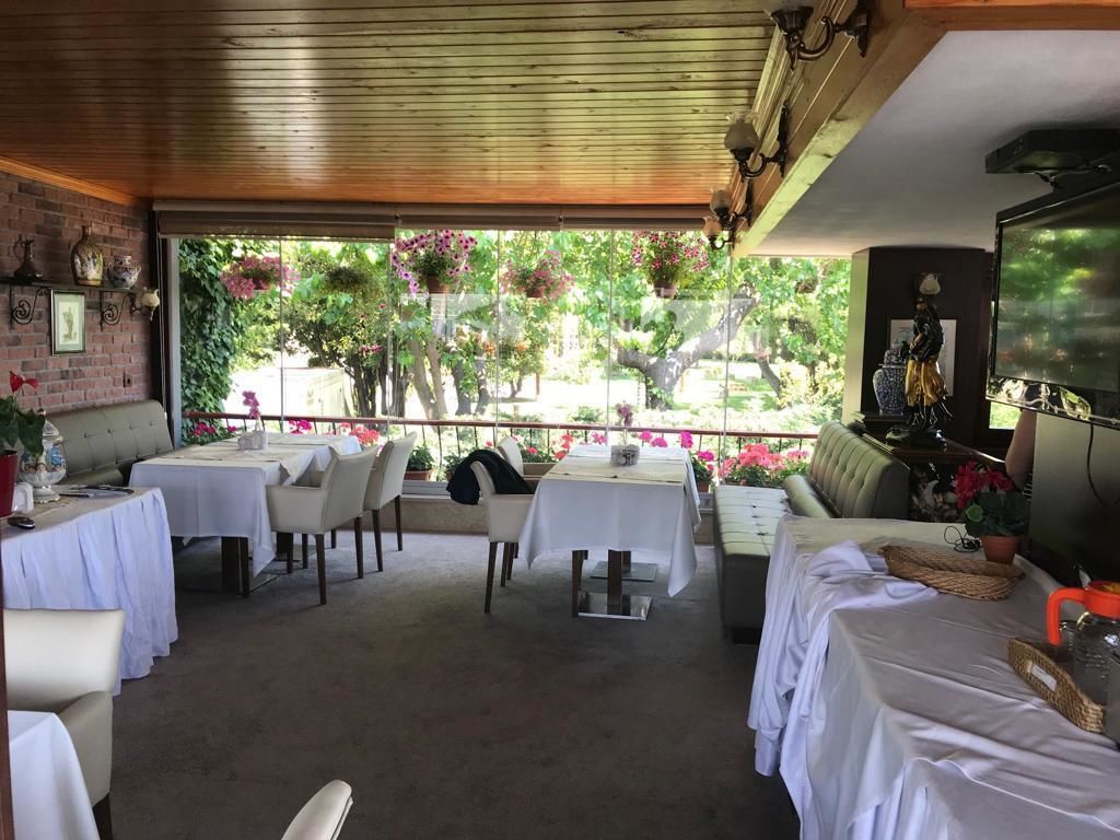 A covered outdoor dining area with tables and chairs, a television, and a view of a garden with trees and flowers.