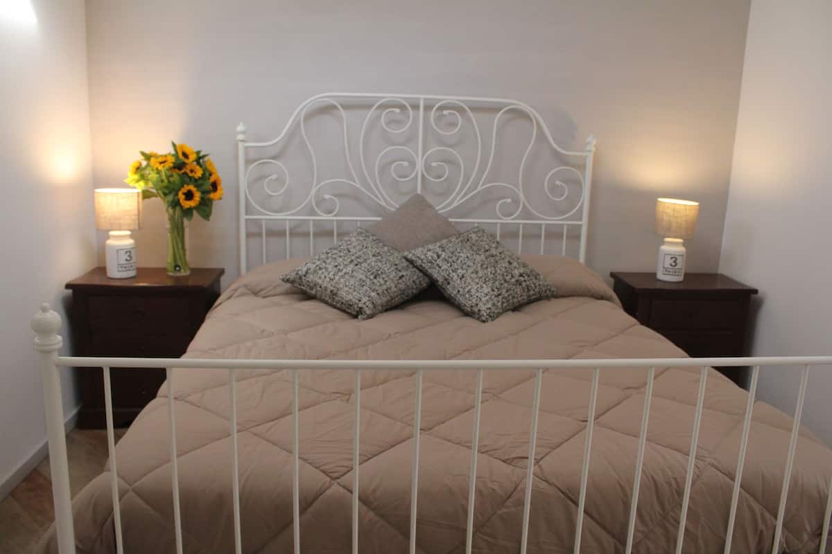 Una cama con cabecera de metal blanco, dos mesitas de noche con lámparas y un jarrón con girasoles.