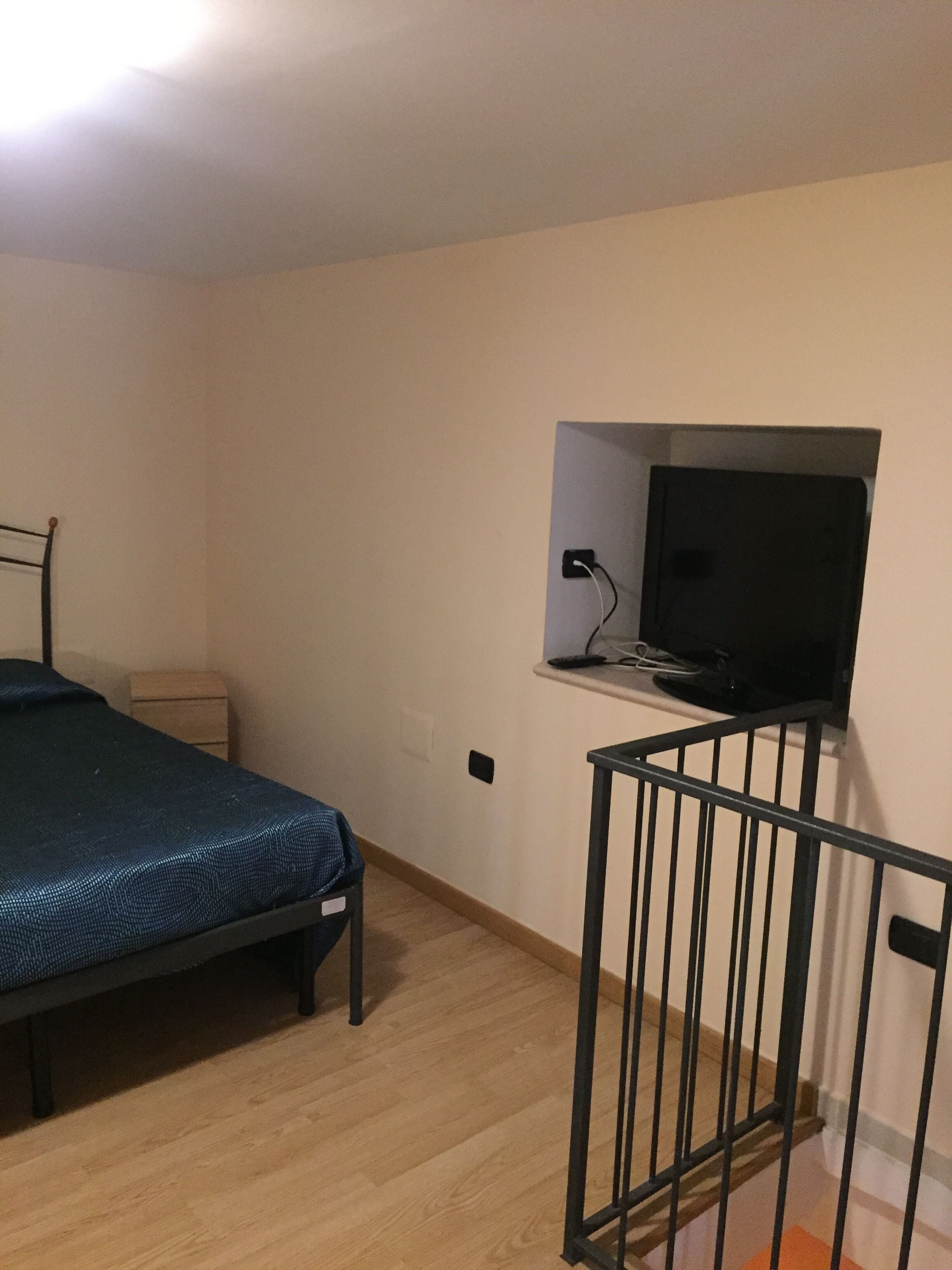A bedroom with a single bed, a small bedside table, a TV mounted on the wall, and a crib in the corner.