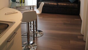 Una sala moderna con un sofá de cuero marrón, una mesa de centro de vidrio y una silla blanca. Hay una zona de cocina con encimera blanca y un lavabo.