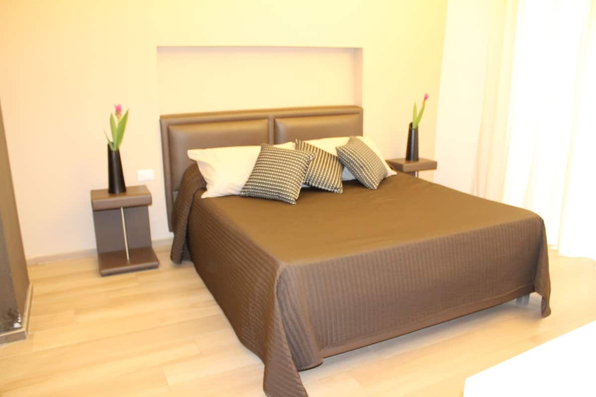 Una cama bien hecha con una colcha marrón y varias almohadas, dos mesitas de noche con jarrones de flores y un piso de madera claro.