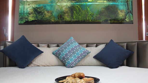 Una cama con sábana blanca, dos almohadas azules y un plato de comida delante.