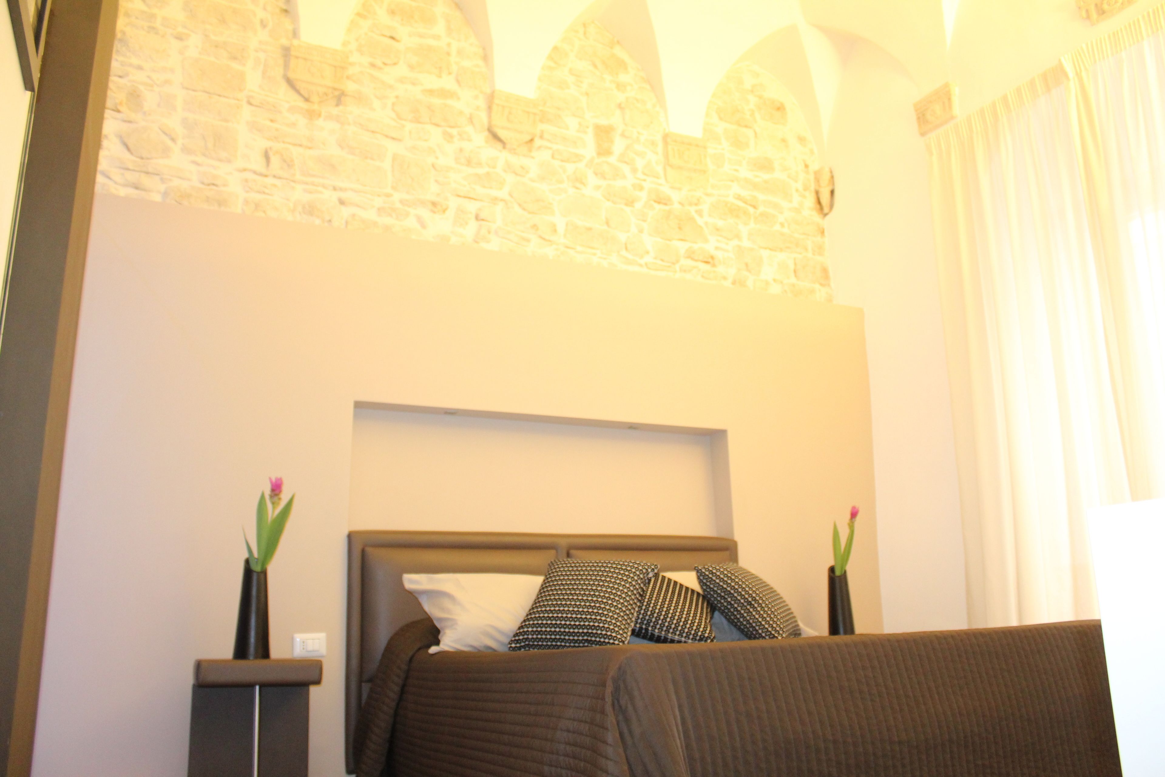 Une chambre d’hôtel avec un mur en pierre, un lit avec une tête de lit, deux tables de chevet avec des vases et une fenêtre avec des rideaux.
