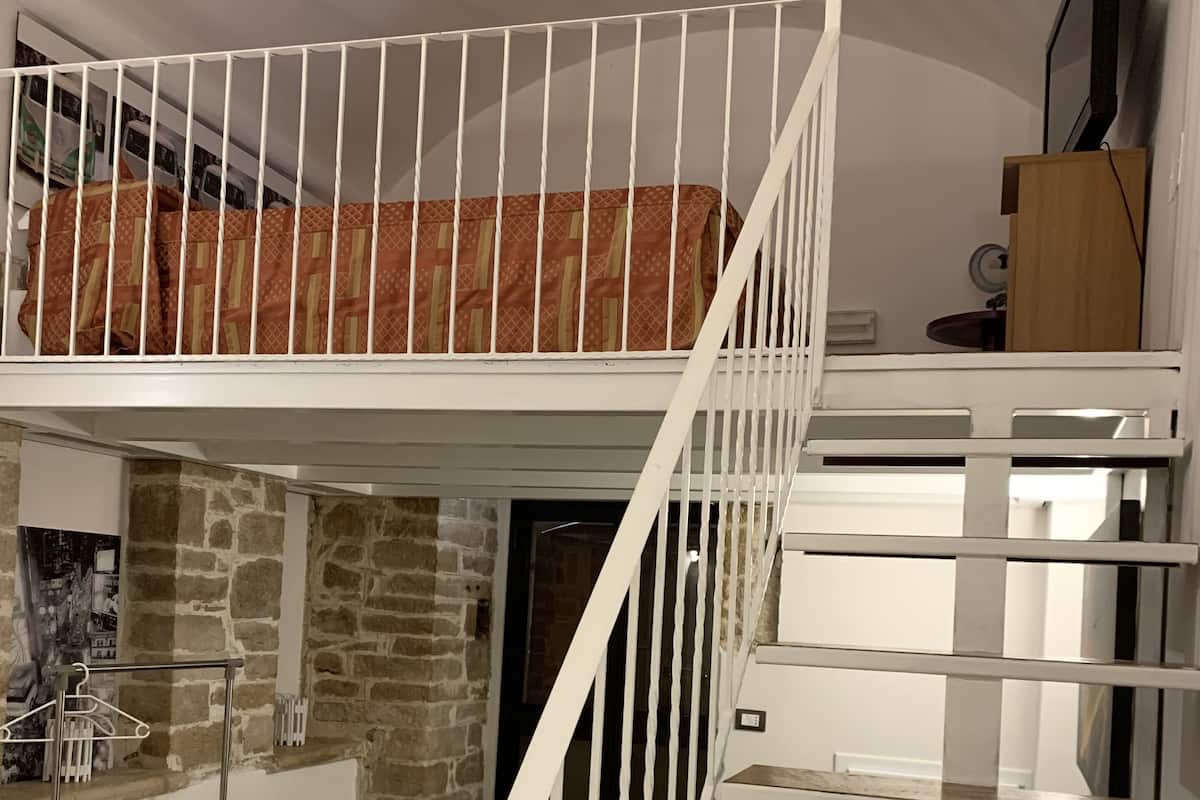 Habitación de varios niveles con una escalera blanca, un altillo con ropa de cama estampada y una pared de piedra.