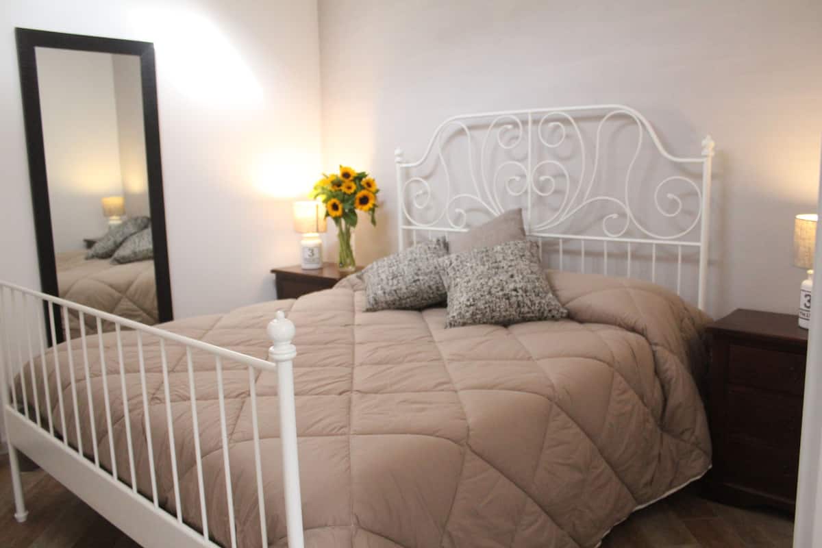 Un dormitorio con cama, mesita de noche, espejo y un jarrón con flores.