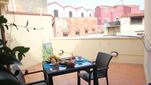 Un balcón con una mesa dispuesta para el desayuno, que incluye frutas y una taza. Hay sillas alrededor de la mesa, y el balcón da a una hilera de edificios.