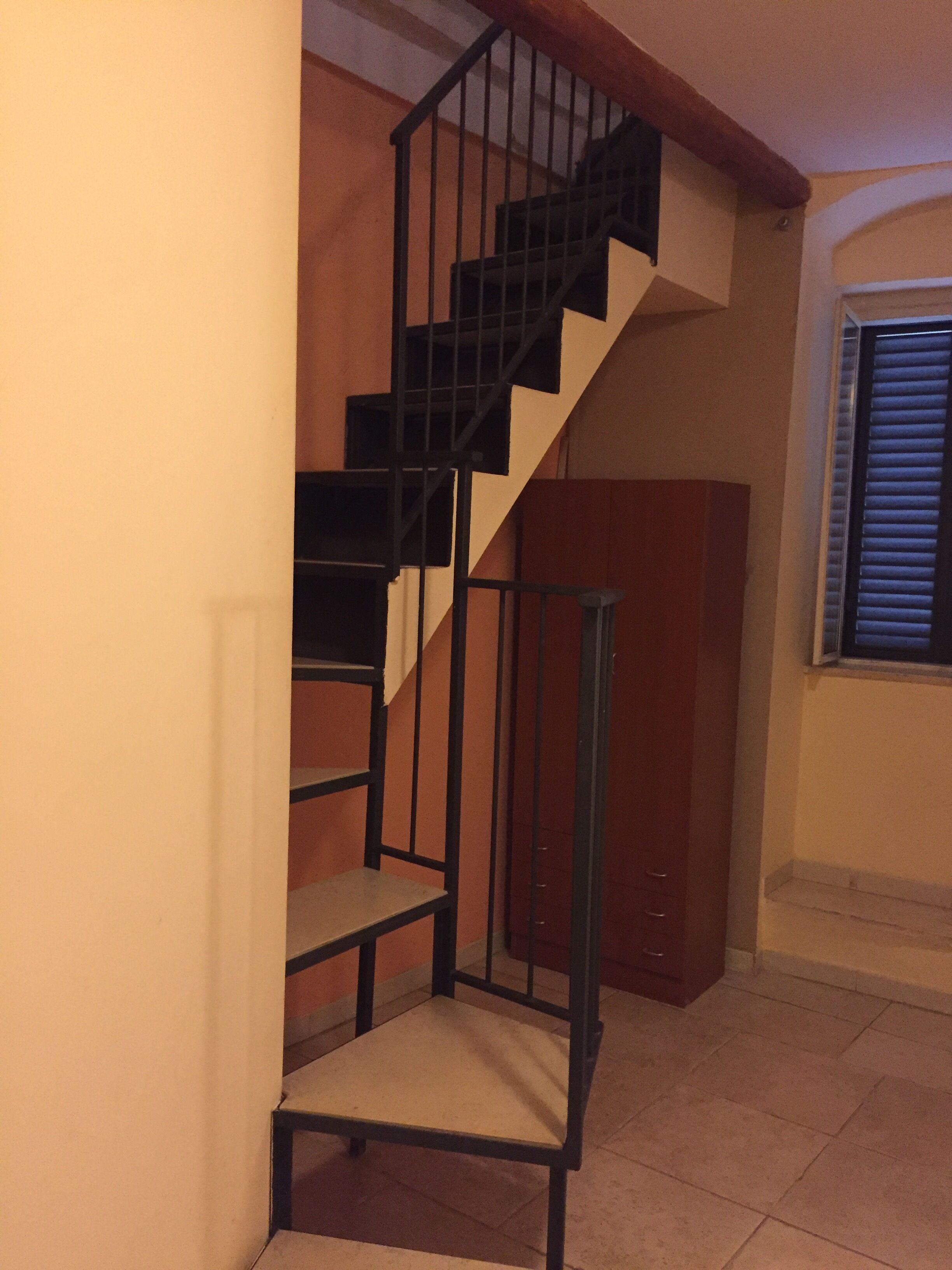 Un escalier avec une chaise en bas, un meuble en bois et une fenêtre avec des stores.
