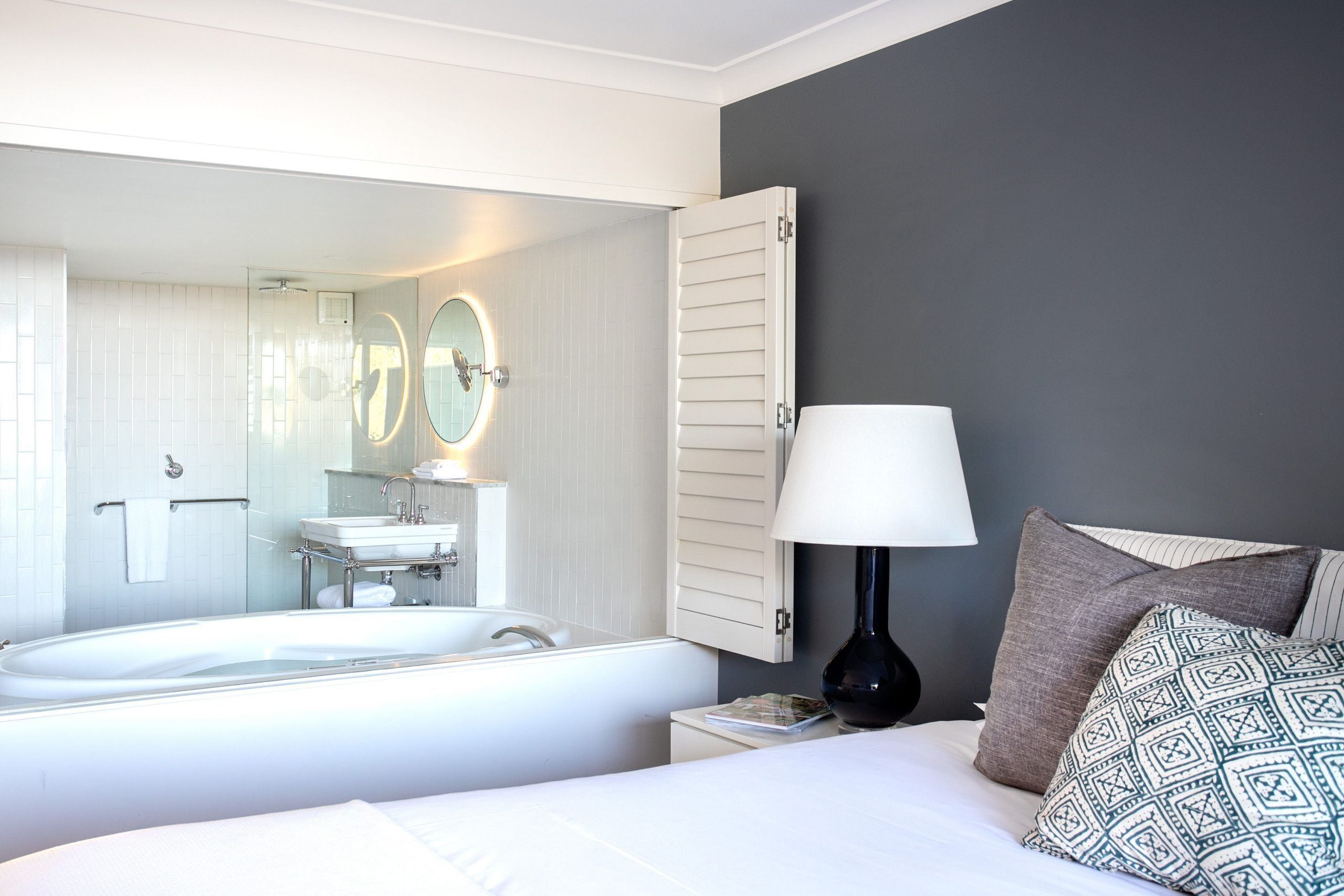 A modern hotel room with a large bed, a bedside table with a lamp, a nightstand, a large mirror, a bathroom with a bathtub, and a towel rack.