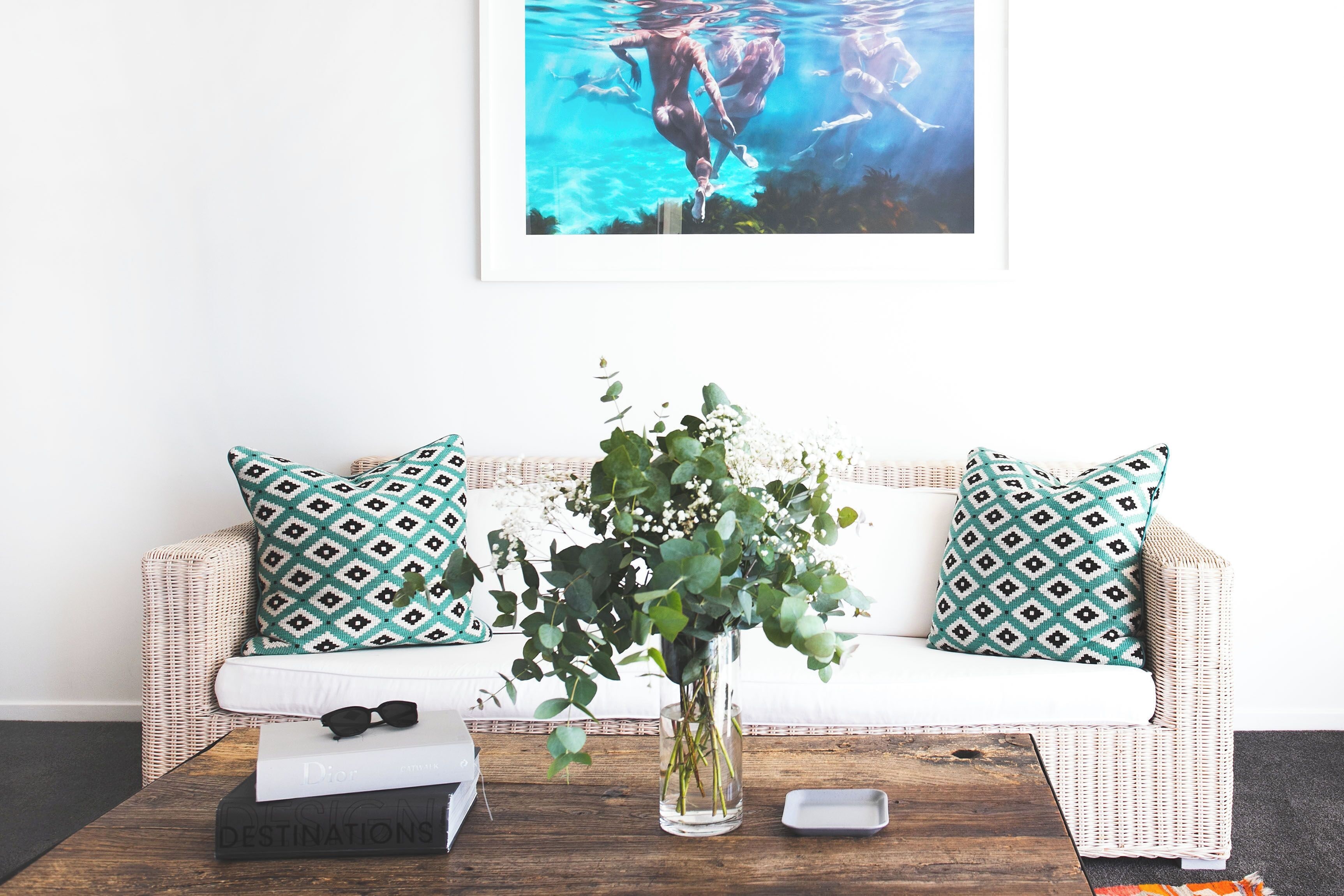 A living room with a white sofa, green patterned pillows, a vase with green foliage, a book titled 'DESTINATIONS', and a framed picture above the sofa.