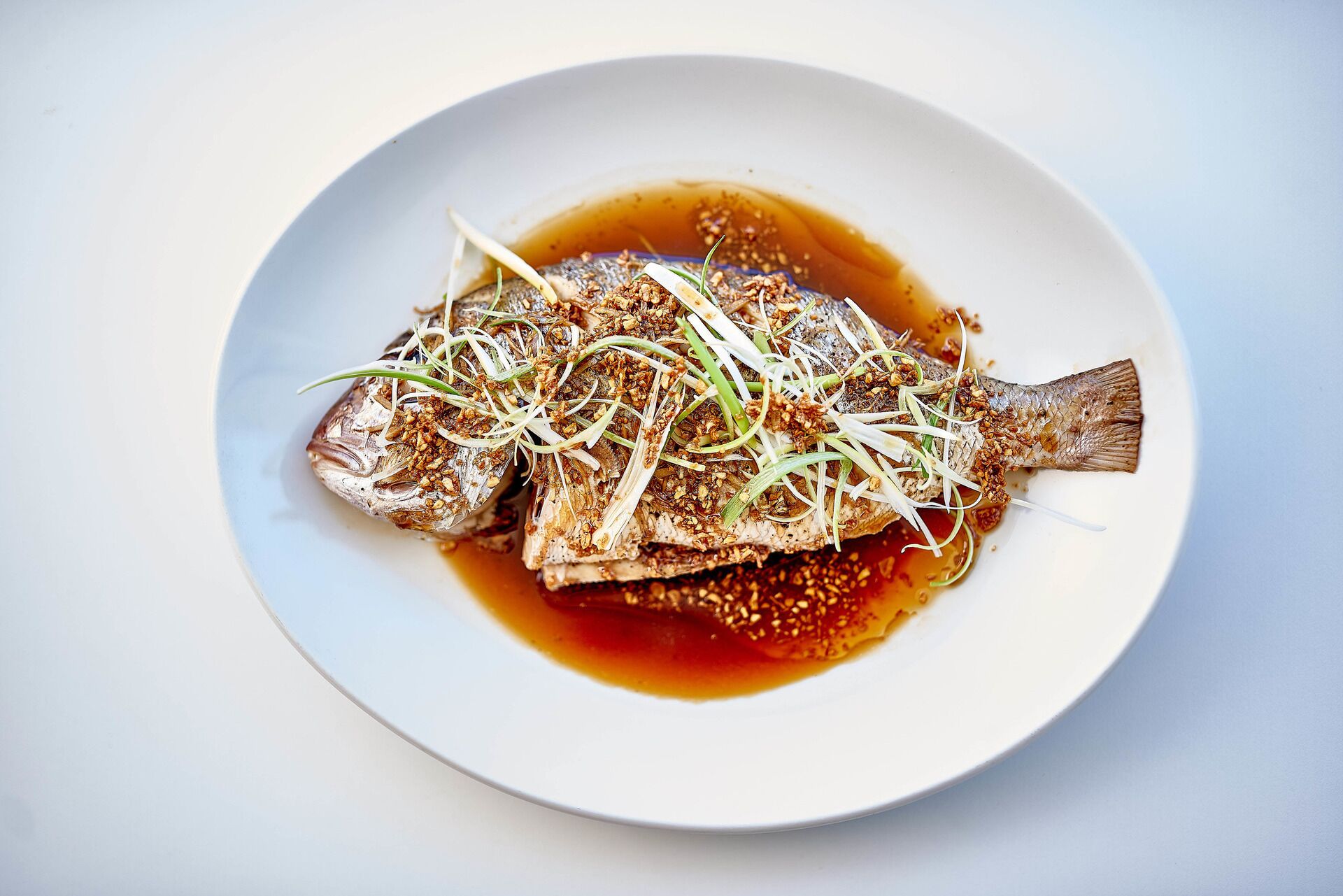 A white plate with a cooked fish garnished with green onions and sesame seeds, served in a dark sauce.