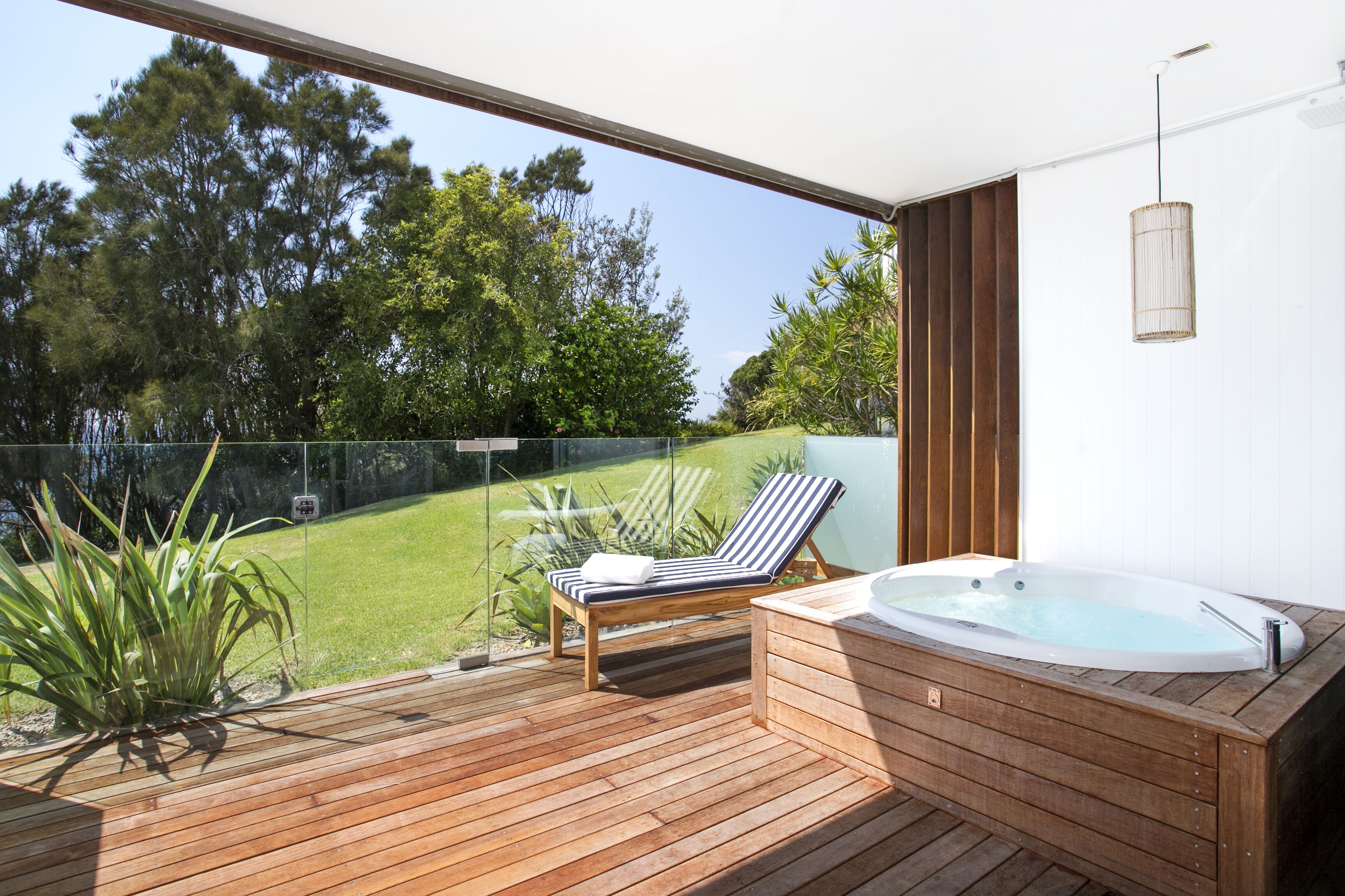 A wooden deck with a hot tub and a lounge chair, overlooking a grassy area with trees.