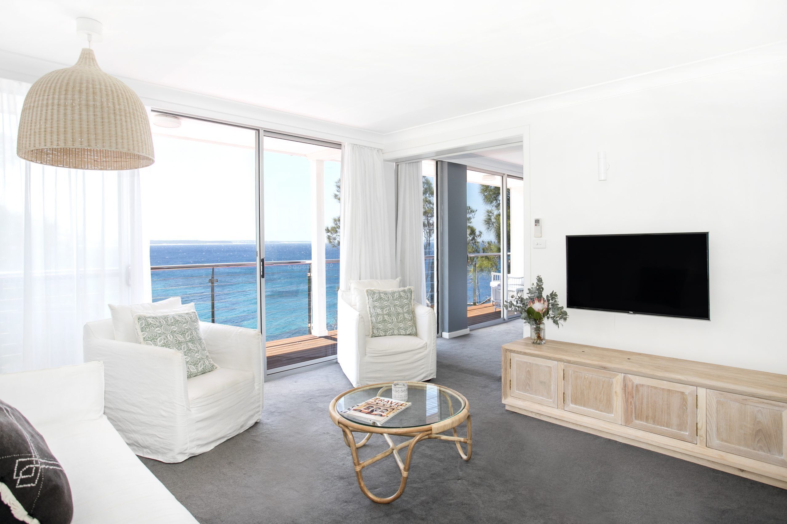 Una sala de estar moderna con un televisor grande, muebles blancos, una mesa de centro de vidrio y vista al océano.