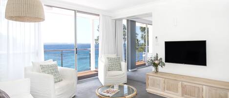 A modern living room with a large TV, white furniture, a glass coffee table, and a view of the ocean.