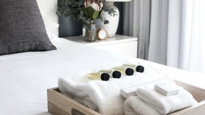 A neatly made bed with white linens, a wooden tray with folded towels, and a small vase with flowers on a bedside table.