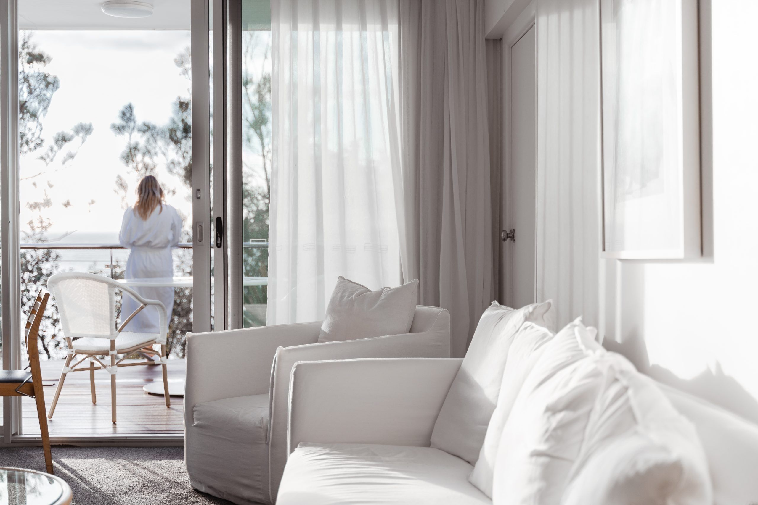 A modern living room with a white sofa, a glass table, and a view of a balcony with a person in a white robe.