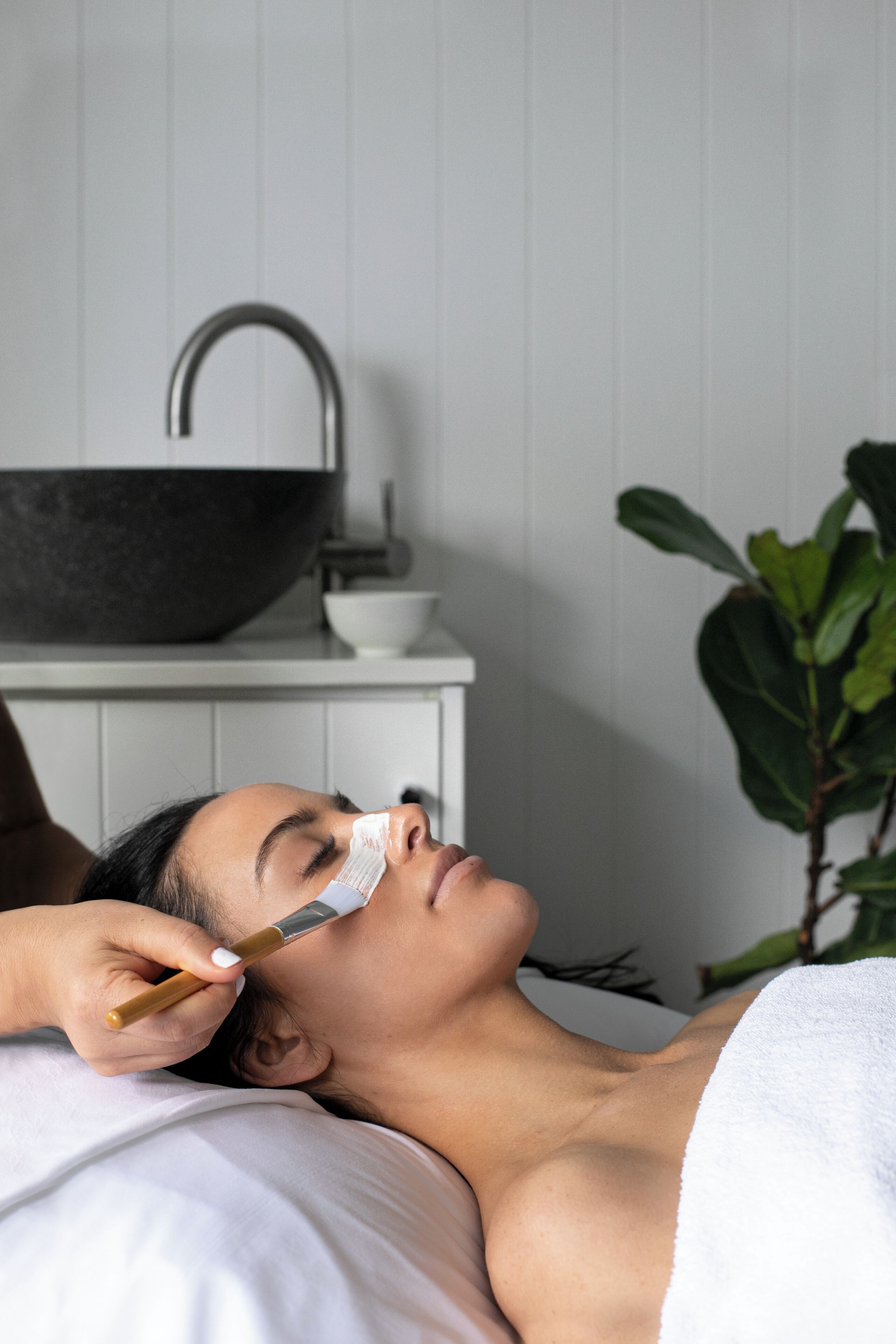 A person lying down with a towel covering their lower body, a sink, and a plant in the background.