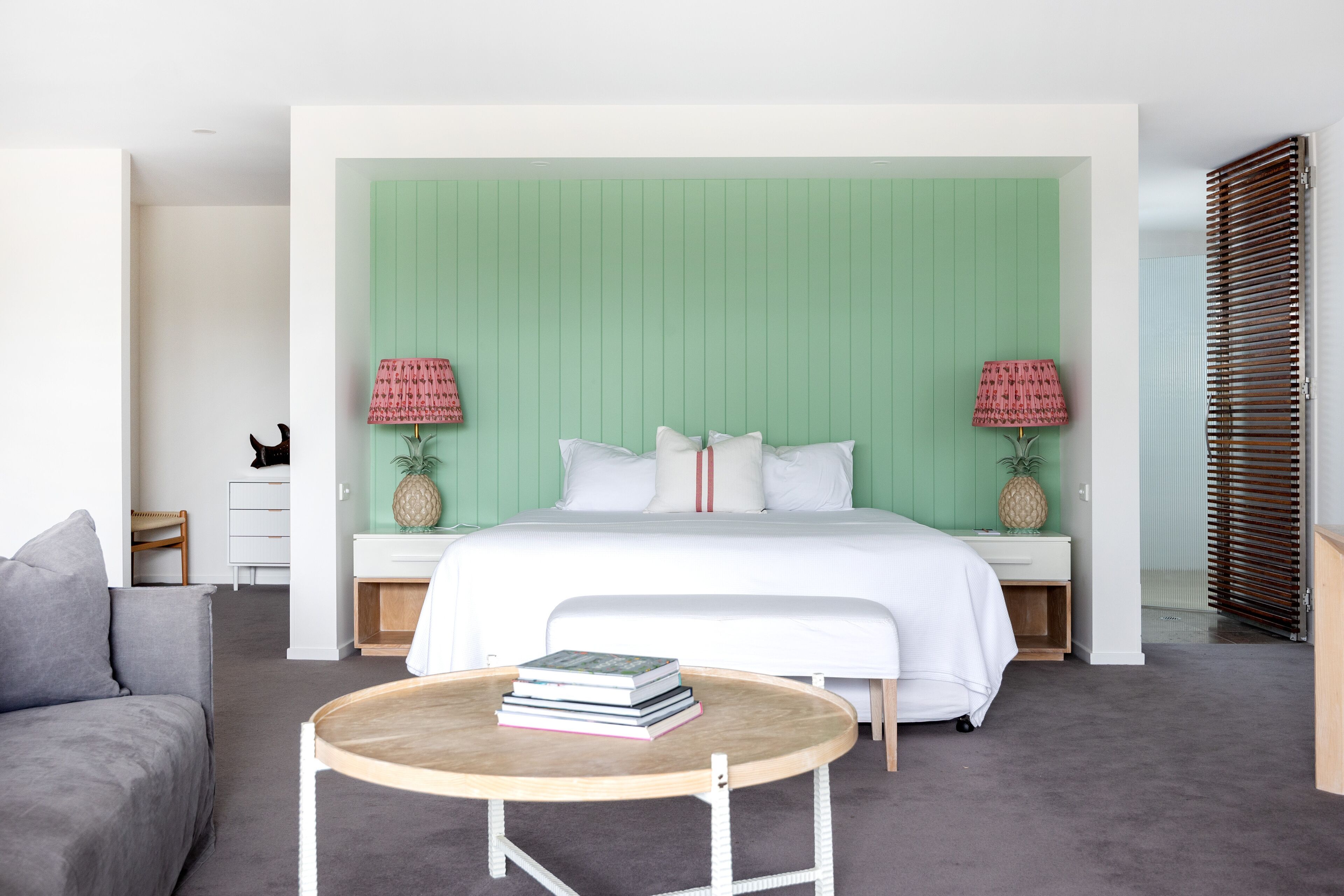 A modern bedroom with a green headboard, a bed with white bedding, two bedside lamps, a round wooden table, and a grey sofa.