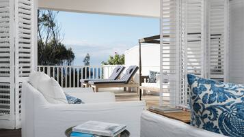 A sunlit outdoor seating area with white furniture and blue patterned cushions, overlooking a wooden deck and trees.