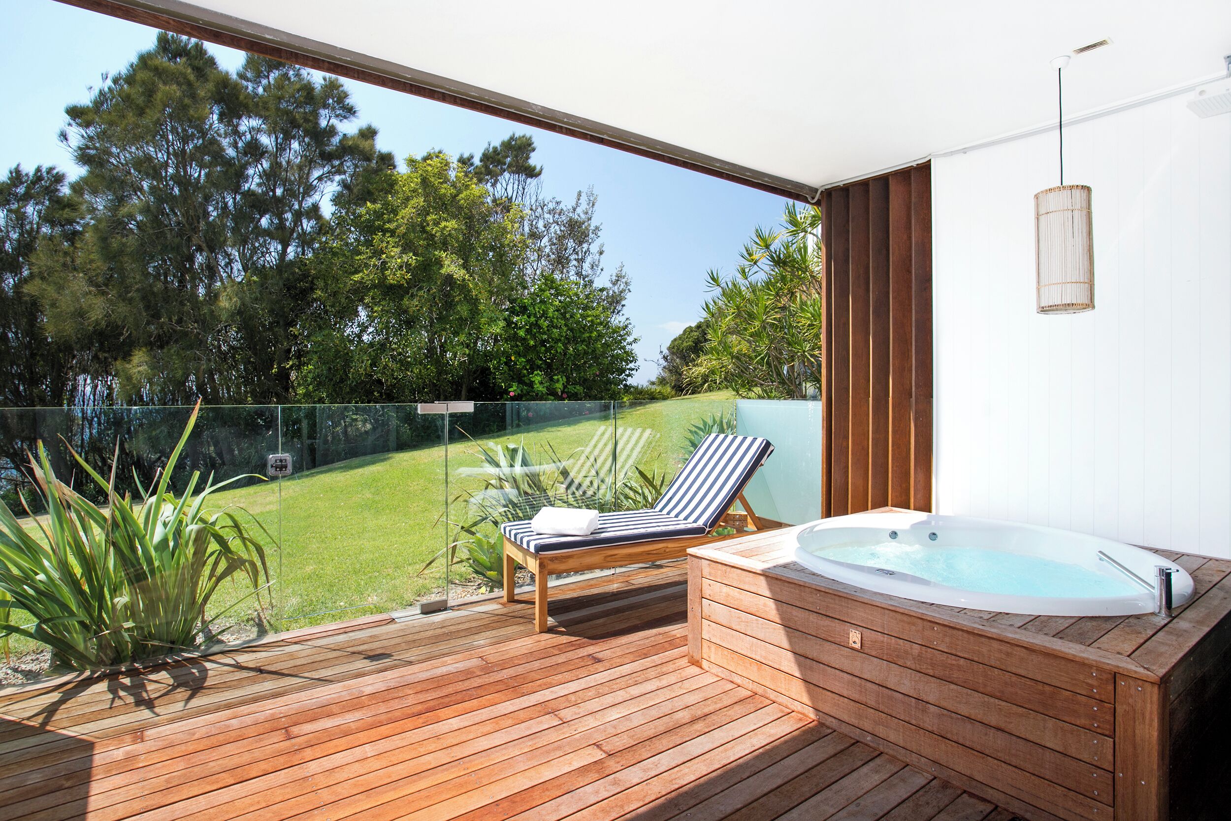 Una terraza de madera con un jacuzzi y una silla de descanso, con vista a una zona verde con árboles.