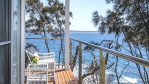 A wooden deck with white chairs and a table, overlooking a coastal area with trees and the sea.