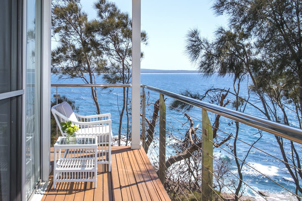 Eine Holzterrasse mit weißen Stühlen und einem Tisch, mit Blick auf eine Küstenlandschaft mit Bäumen und dem Meer.