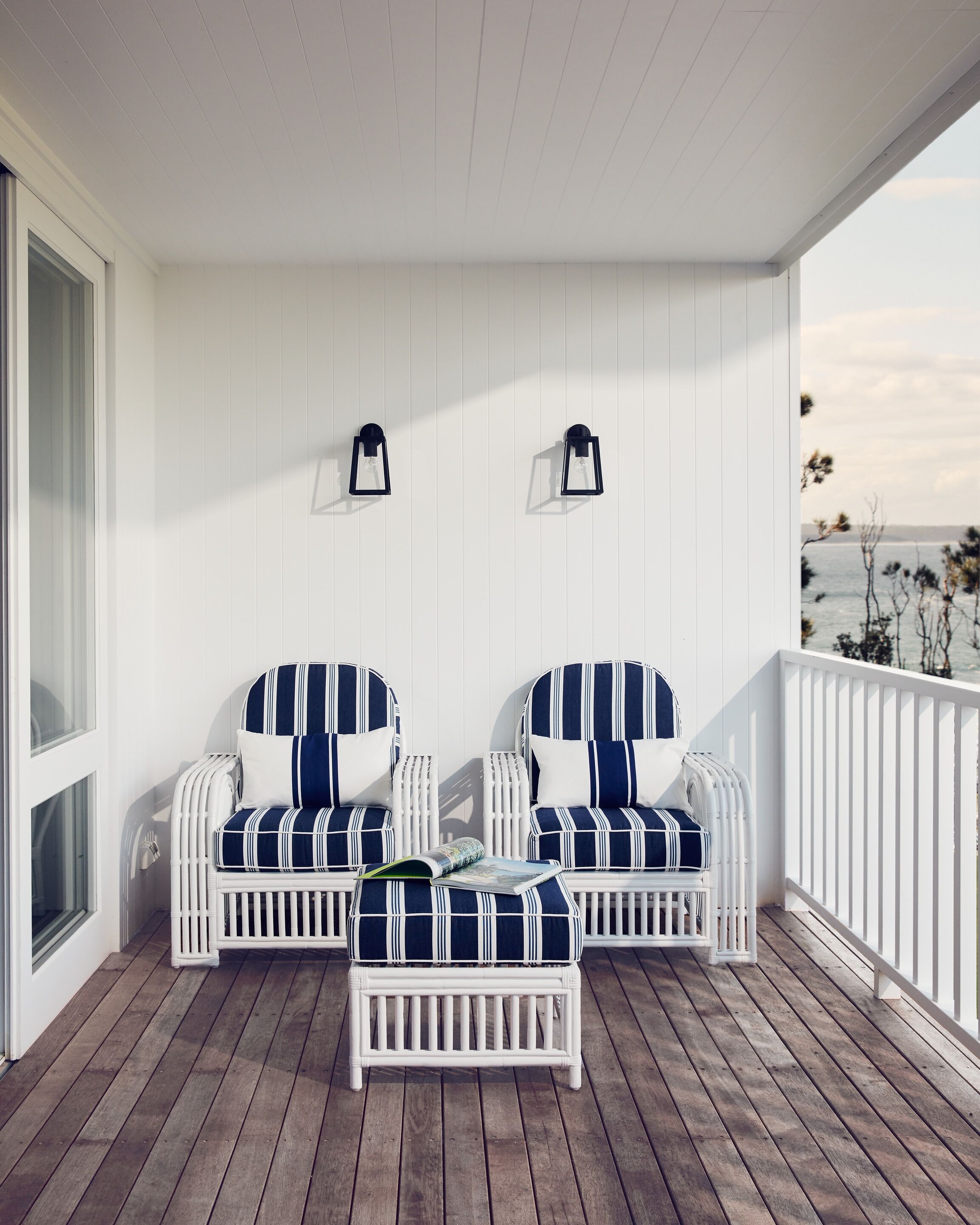 Eine weiße Veranda mit zwei gestreiften Stühlen, einem kleinen Tisch und einem weißen Geländer, mit Blick auf das Meer.