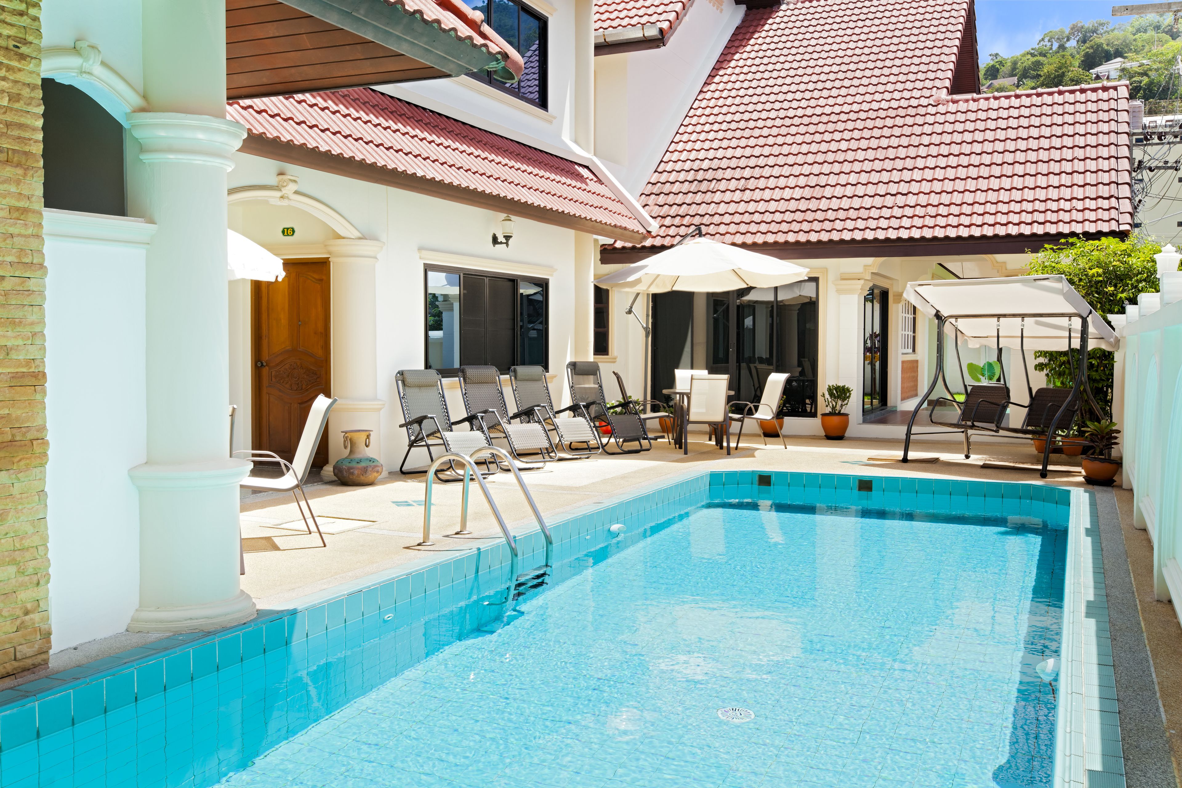 A swimming pool with lounge chairs and umbrellas, surrounded by a building with a tiled roof and a door marked with the number 18.