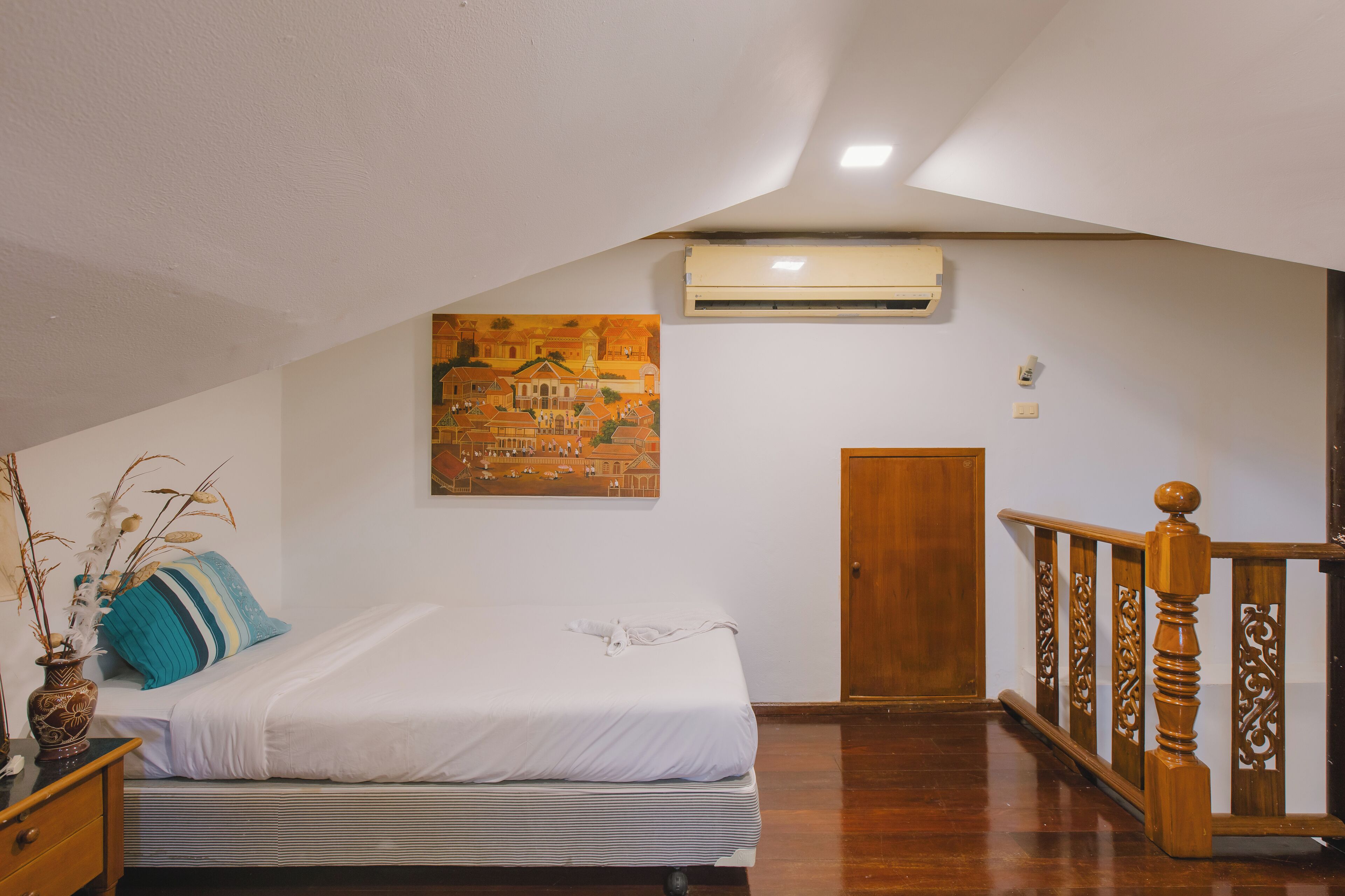 Une chambre à coucher comprenant un lit, une table de chevet, un vase avec des plantes séchées, un climatiseur mural, une porte en bois et un escalier avec une rampe en bois.