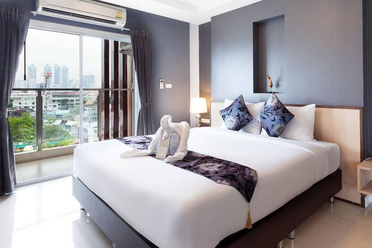 A modern hotel room with a large bed, a bedside table with a phone and a small lamp, and a view of the city through a large window.