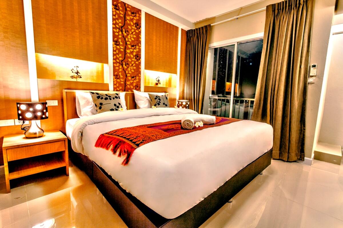 A spacious hotel room with a large bed, a nightstand, a lamp, and a view of the outside through a window with curtains.