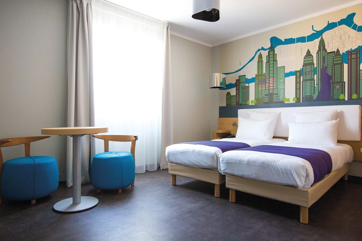 A hotel room with a bed, two blue armchairs, a small table, and a cityscape mural on the wall.