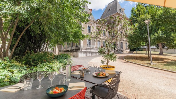 An outdoor dining area with a table set for two, surrounded by greenery and a view of a historic building. - Appart'City Confort Lyon Cité Internationale (Caluire-et-Cuire)