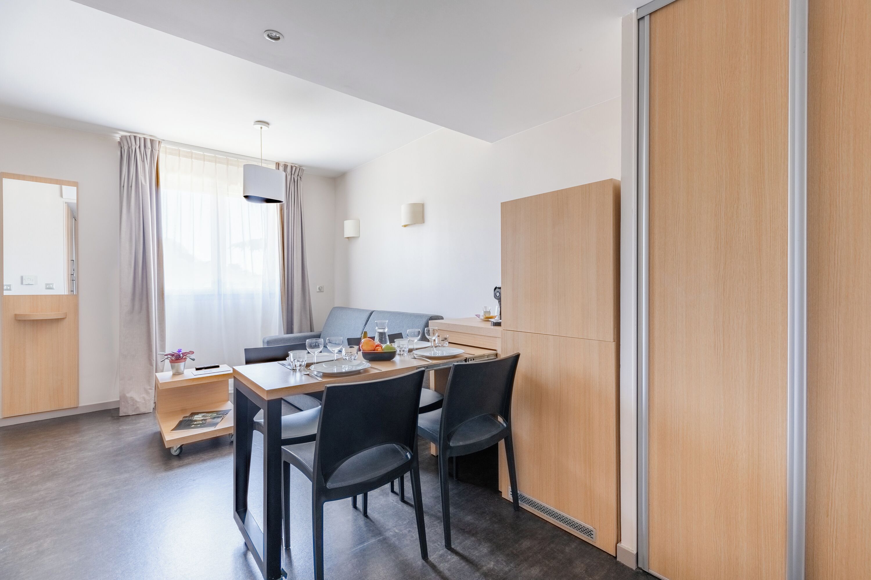 A modern hotel room with a dining area, a wooden wardrobe, and a large window with curtains.
